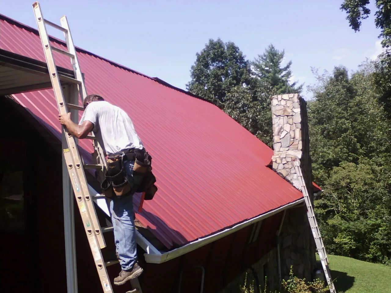Roofer on a ladder beside a red metal roof and stone chimney — Soffit & Fascia Repair by Roofing Brothers in Hansville